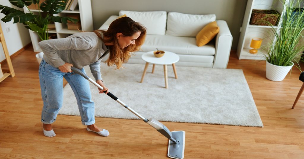 A young woman mops the hardwood floors in a living room with a small couch, table, rug, bookshelves, and plants.