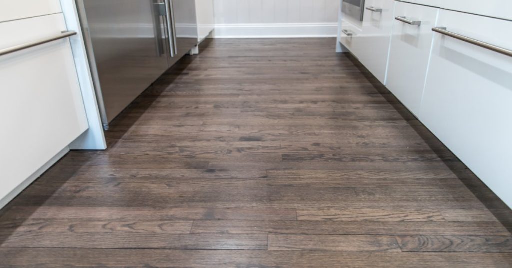 Dark brownish-grey-toned vinyl wood flooring in a small galley-style kitchen space with white cabinetry.